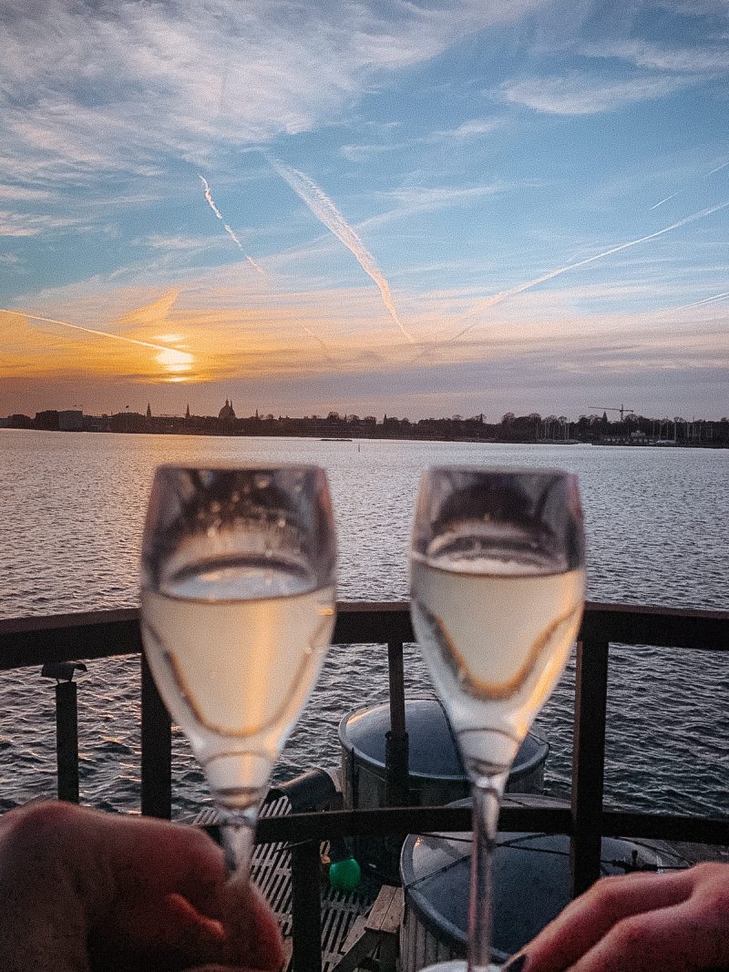 The sun setting in the background, near the sea and a picture two glass of champagne. Things to do in Copenhagen