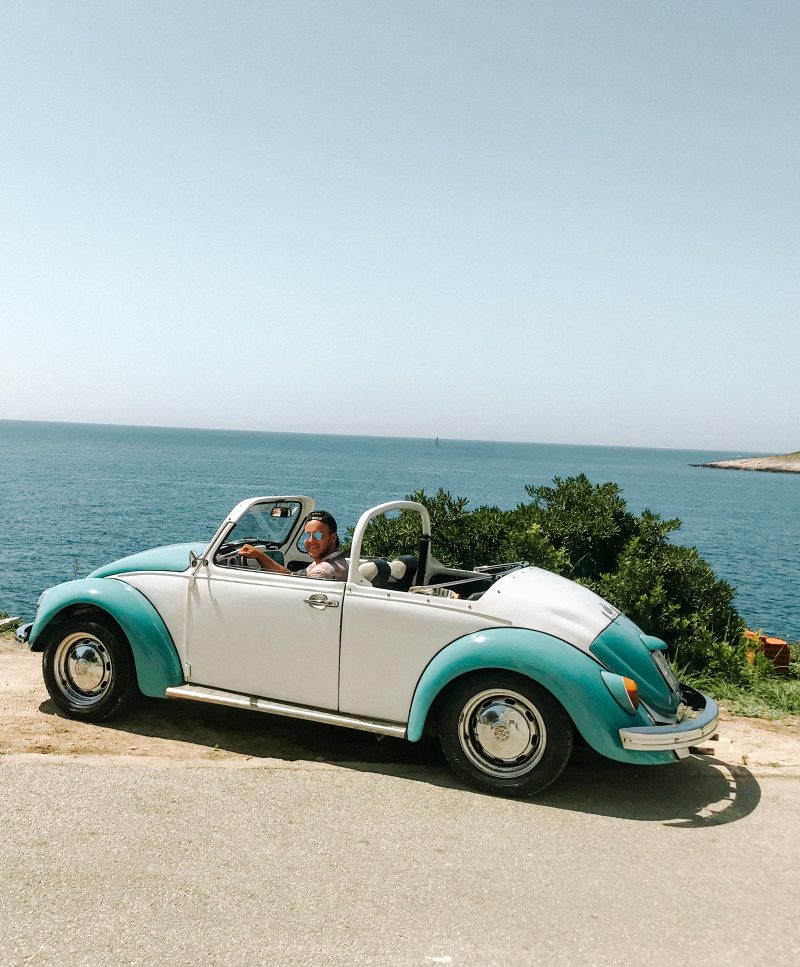 A man in a 60's white and blue VW convertible with the sea in the background. Things to do in Hvar