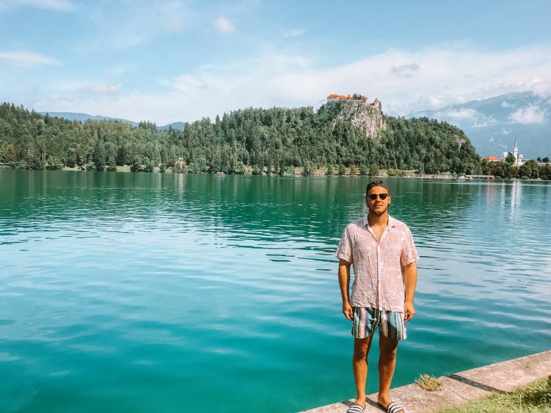A man stood by the bank of the lake with a castle in the background in the hills. Things to do at Lake Bled