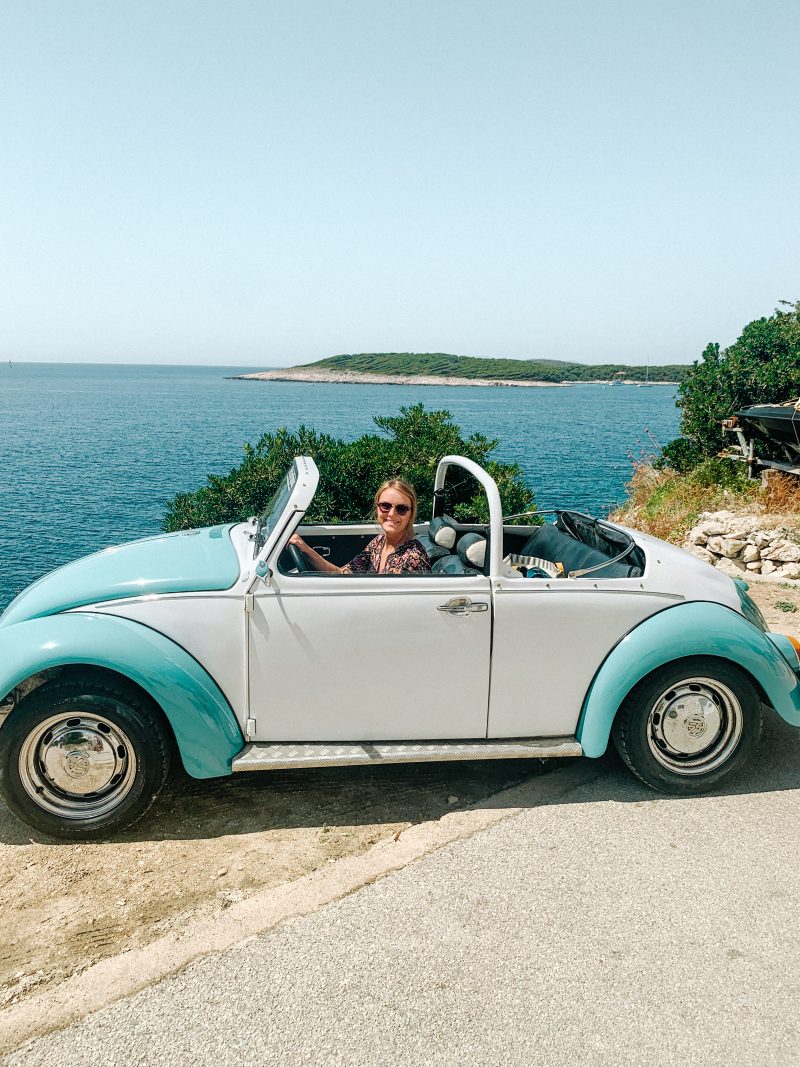 A woman in a 60's white and blue VW convertible with the sea in the background. What to do in Hvar