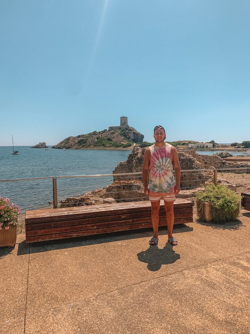 A man stood near the ancient Nora ruins with a castle and ocean in the background. Sardinia beaches