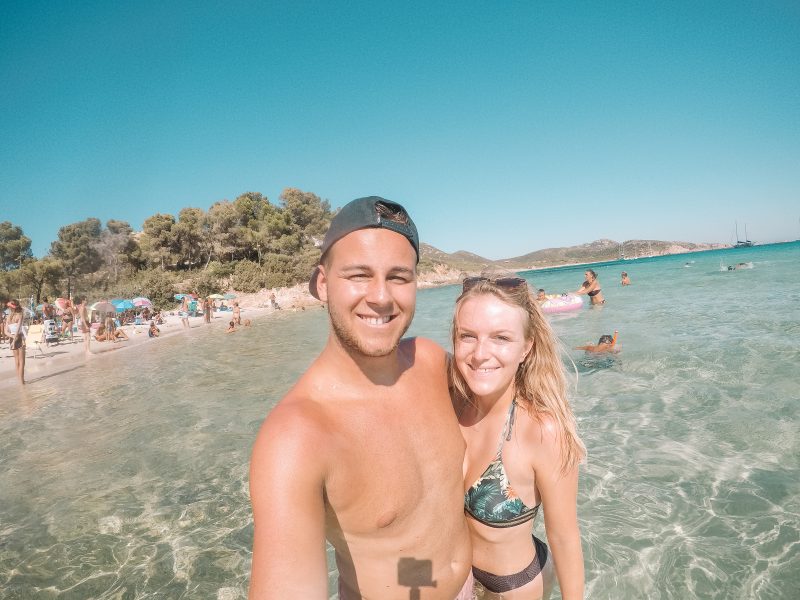 What to din Sardinia. Visit Tuerredda beach. A young couple take a selfie with trees behind them
