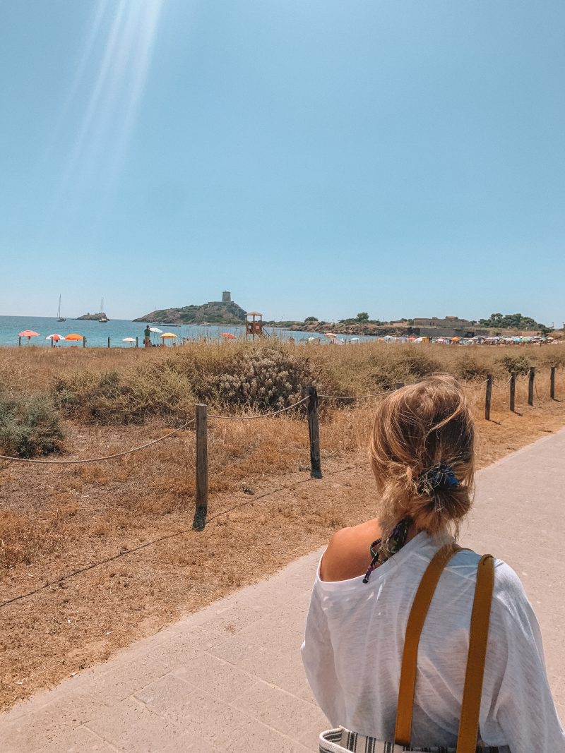 A woman walking up a path near the sea with a castle in the background. Things to do in Sardinia.
