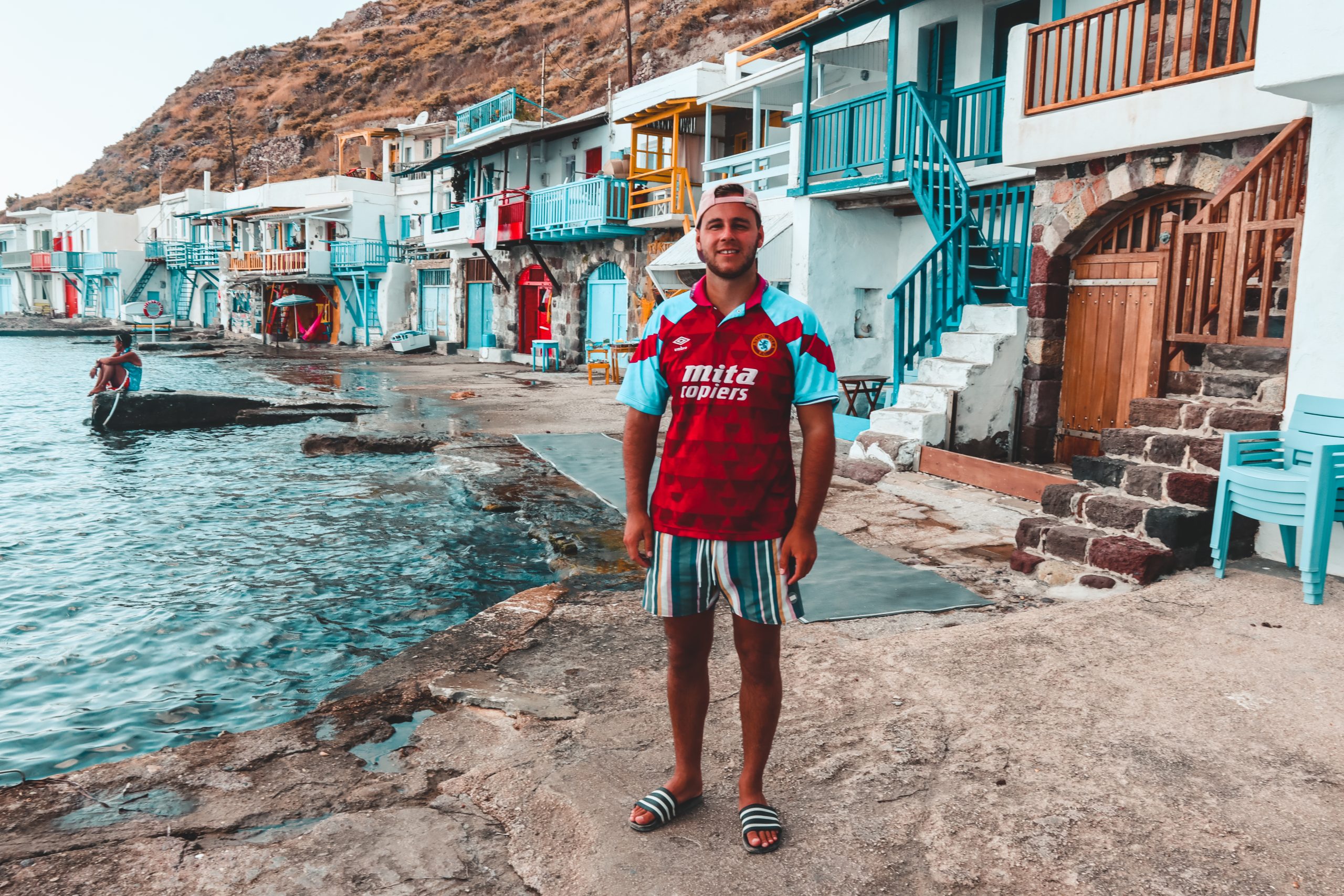 A man stood in front of colourful fishing boat houses. Milos travel guide.