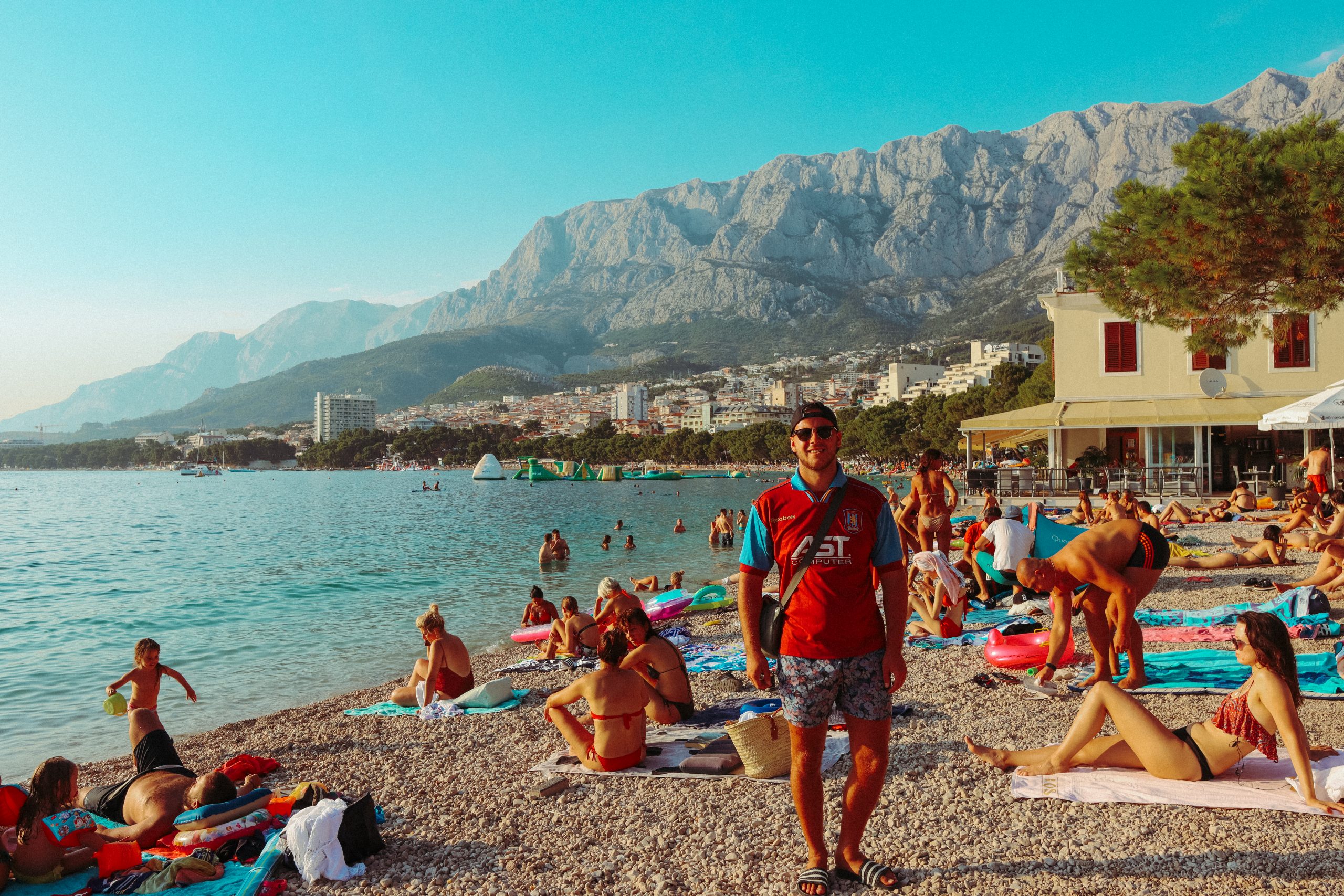 A man stood on a busy pebble beach with Mount Biokovo in the background. Things to see in Makarska