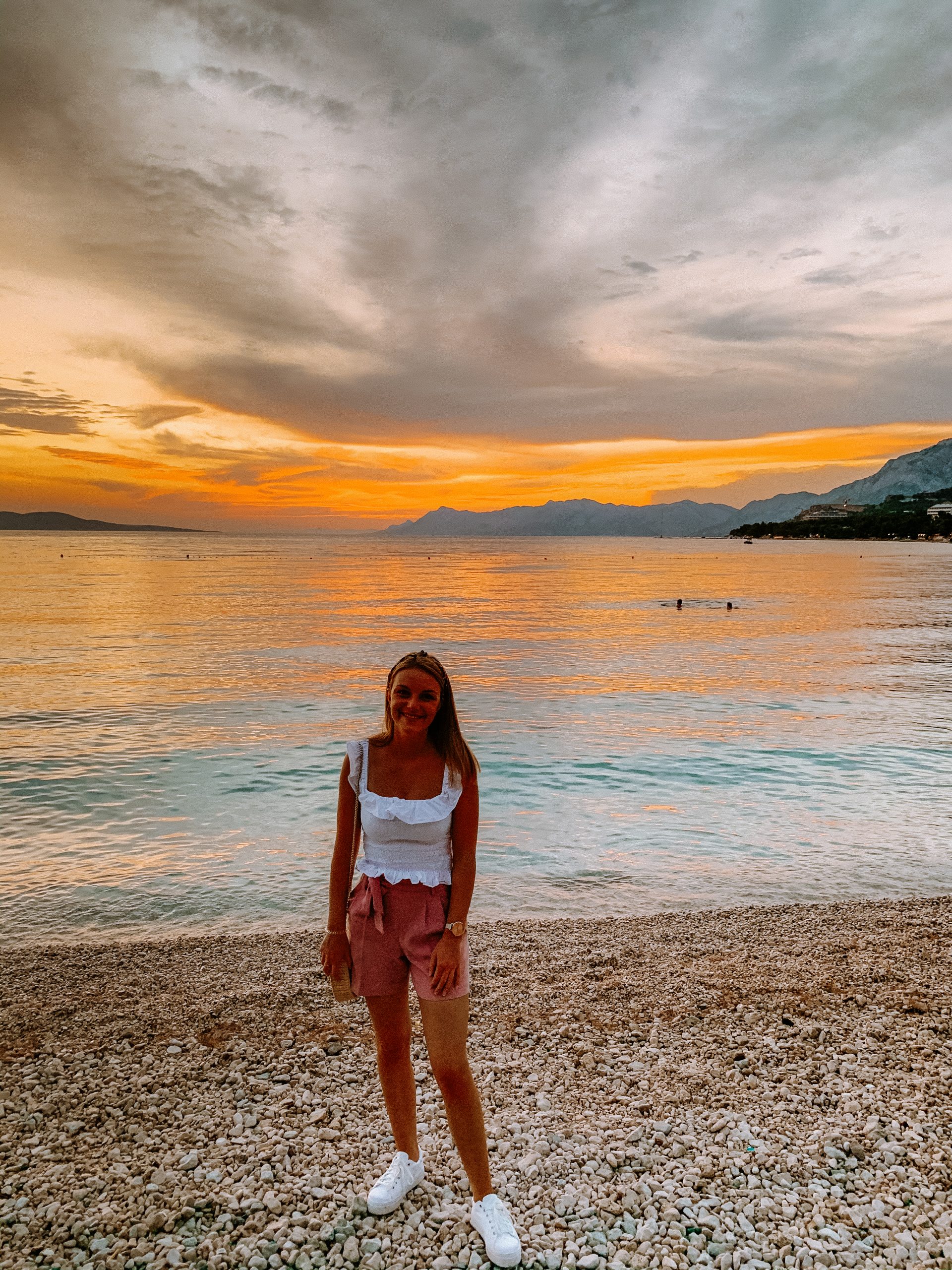 A woman stood on a pebbly beach with the sunset in the background