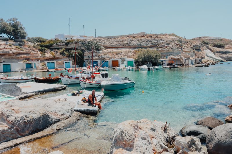 A man sat next to boats and colourful boathouses in Milos