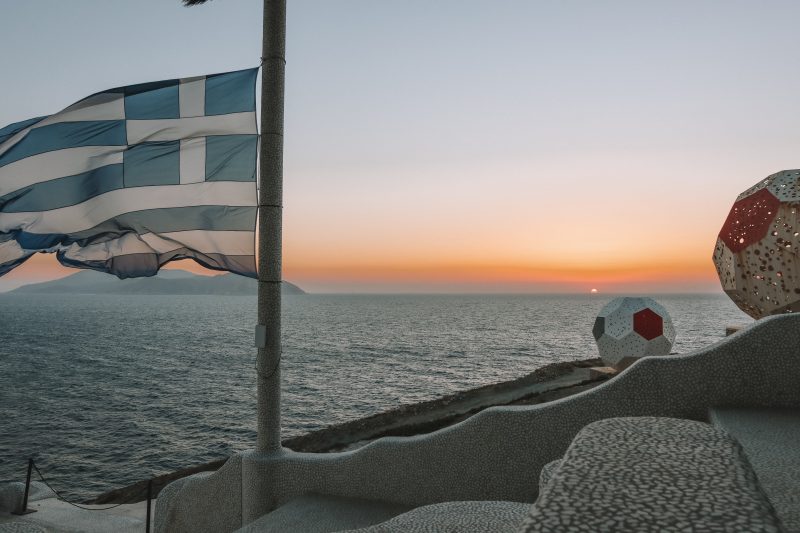 A greek flag with the sun setting in the background. Ios travel blog, pathos lounge