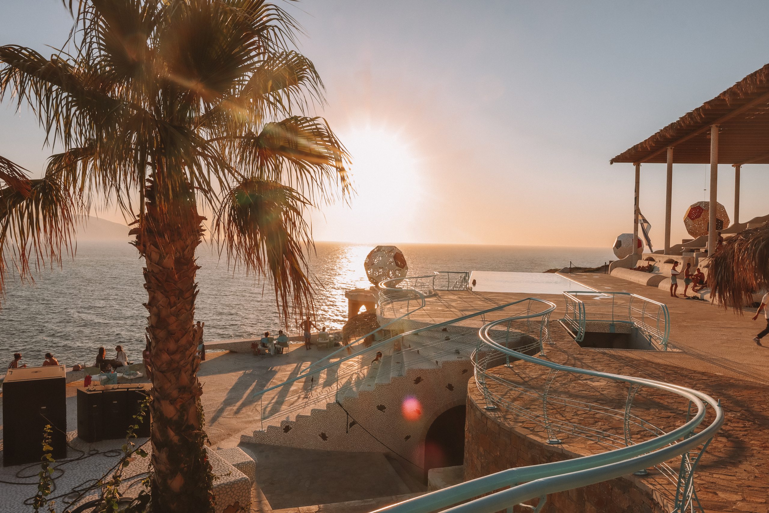 A palm tree and a beach club with the sun setting in the background. Things to see in Ios, Pathos lounge