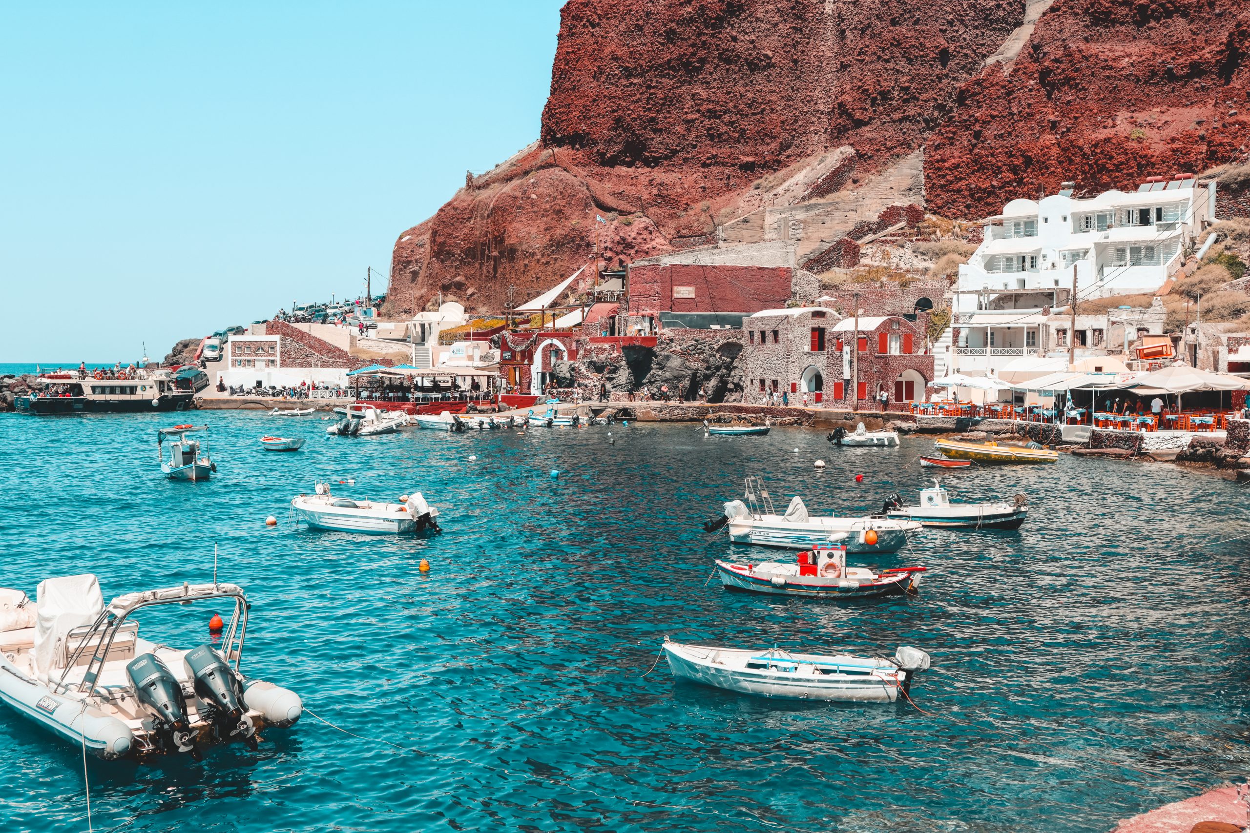 Boats docked by Ammoudi Bay with red cliffs in the background.