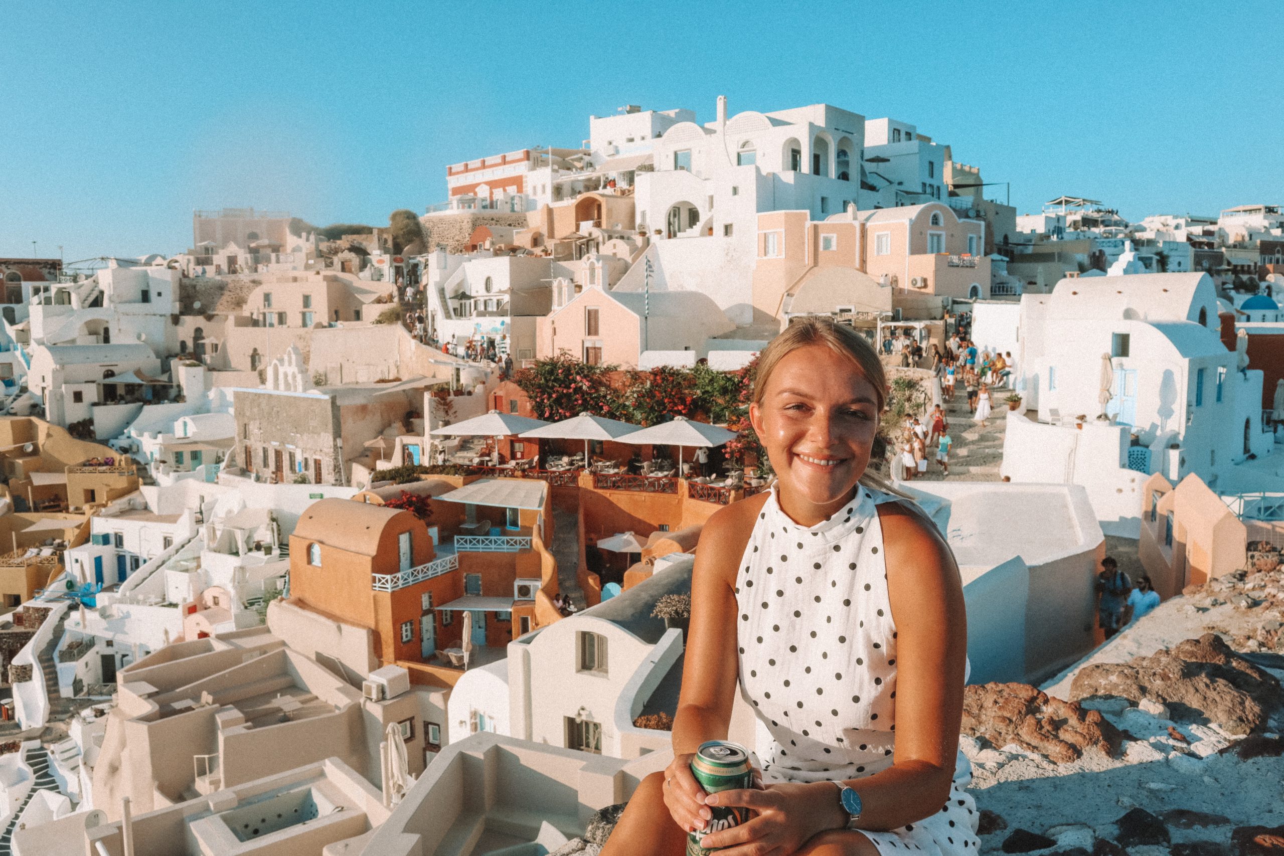 A woman sat with a can of Mythos and pastel buildings in the background. Things to do in Oia