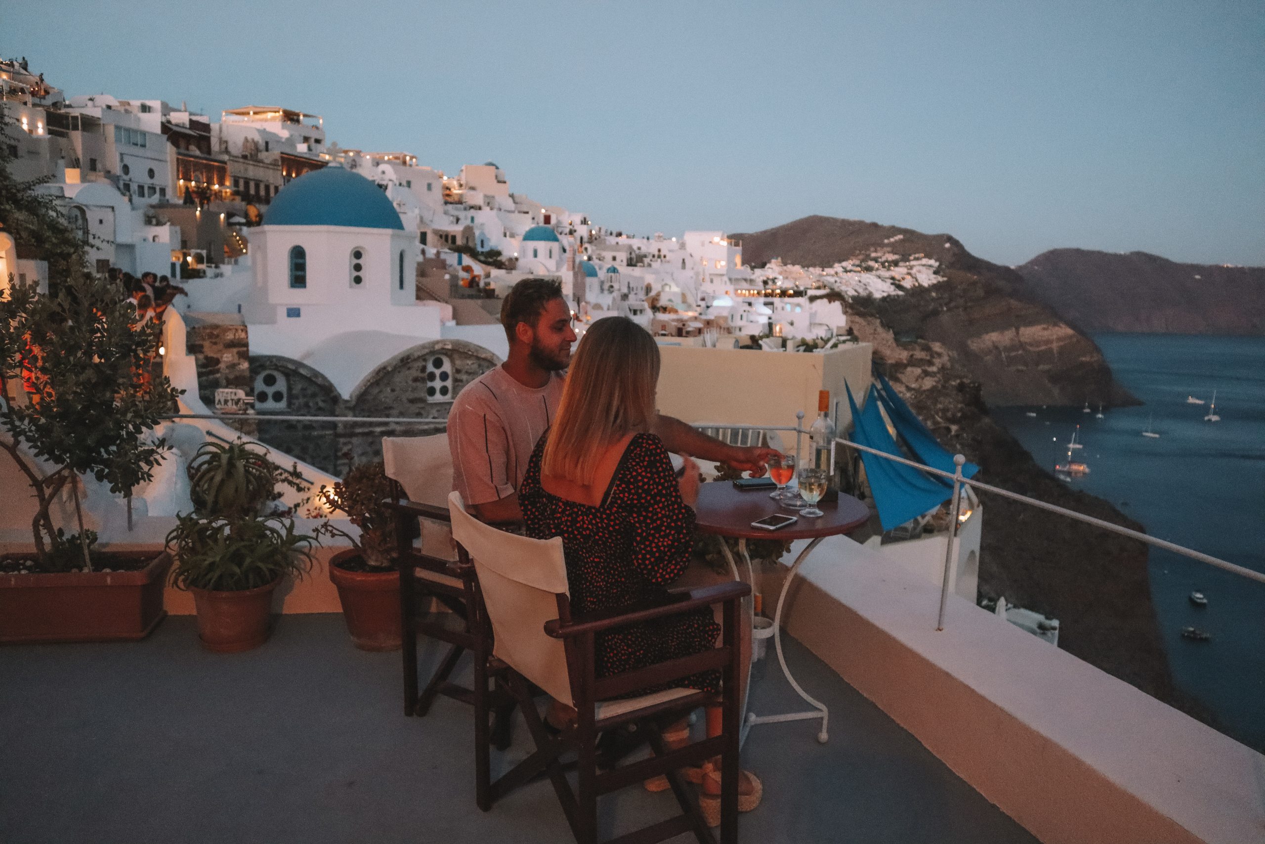 A couple drinking wine on their balcony in the evening with blue domed buildings in the background in Oia