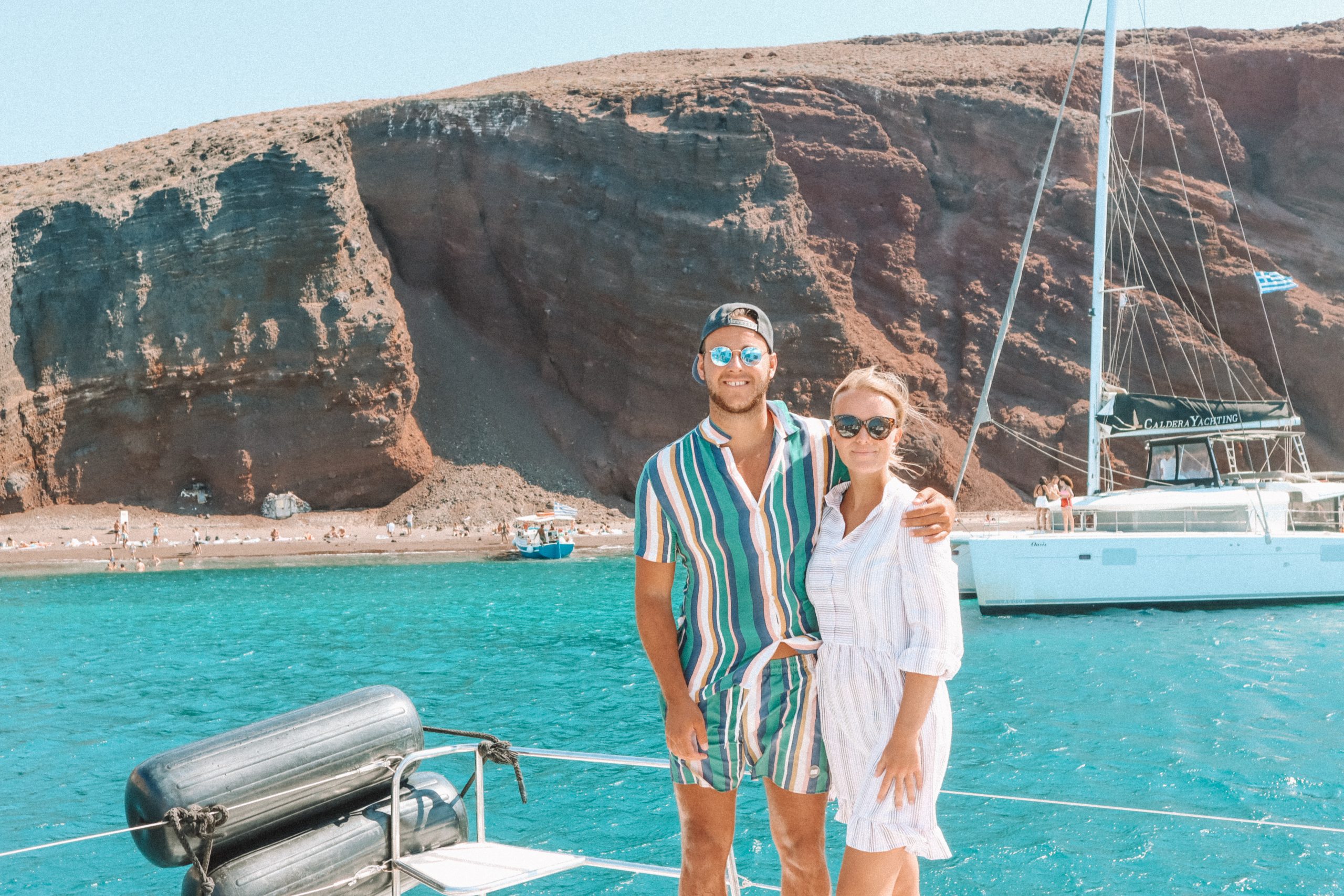 A couple stood on a boat with turquoise water and the red beach in the background. Things to do in Santorini