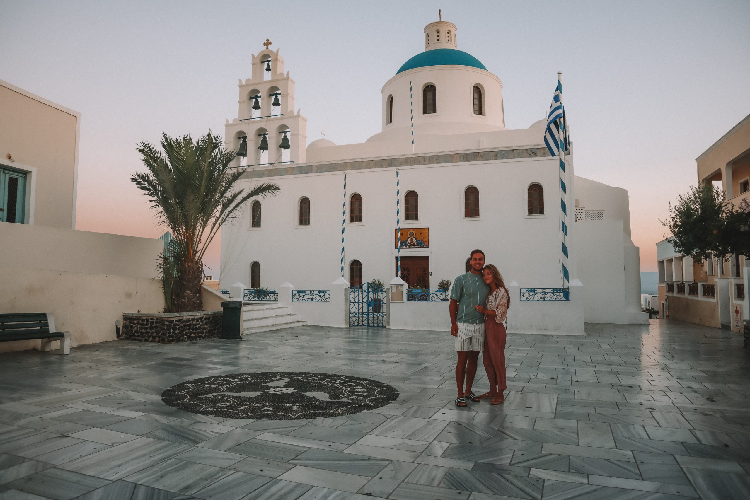 A couple stood in front of a blue domed building and bells during sunrise. Things to do in Santorini