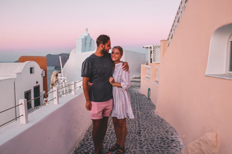 A couple stood next to pastel coloured buildings and a pink sky at sunrise.