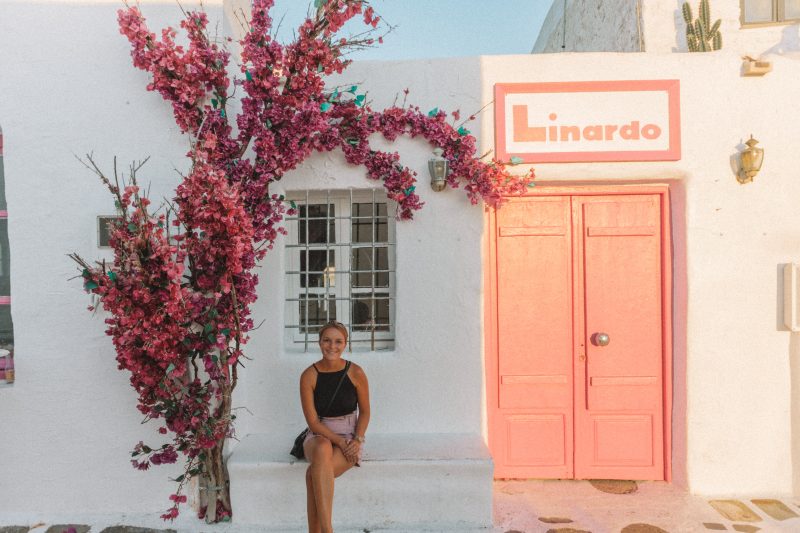 A woman sat on a white ledge next to pink flowers and a pink door in Naoussa.