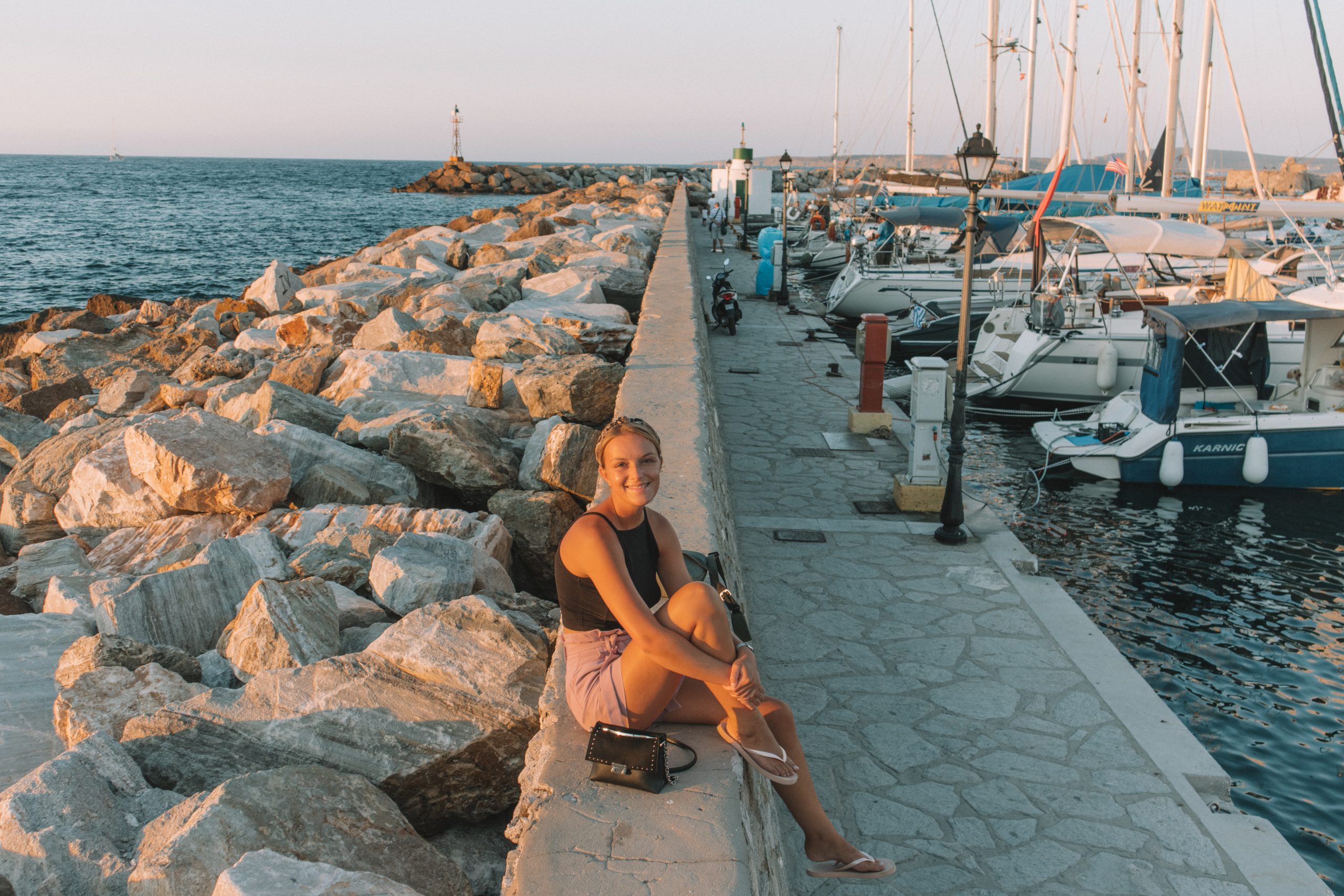 A woman next to the rocks and harbour of Naoussa during sunset.
