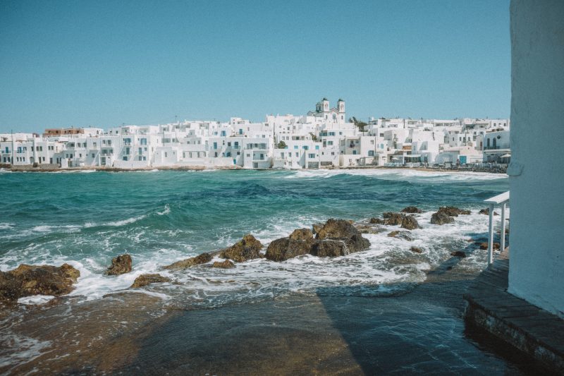 The sea and rocks with whitewashed buildings in the background of Naoussa. Paros travel blog